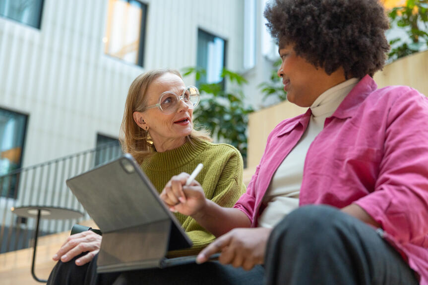 Two women are sitting on stairs, talking. One of them has a tablet in her lap.