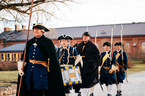 Fem män i mörka historiska dräkter går på gator i Sveaborg.